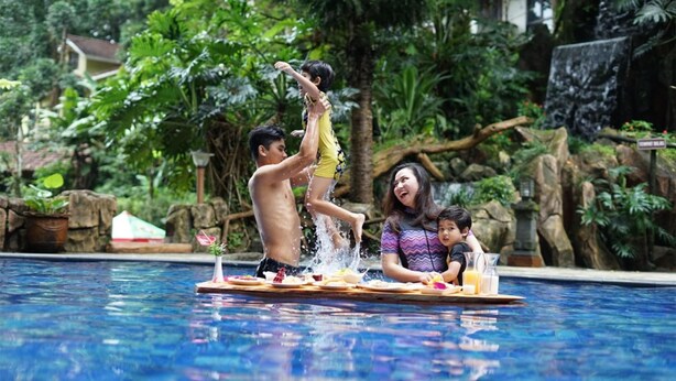 A swimming pool with a wooden deck, surrounded by lush greenery and a waterfall. - Safari Lodge Hotel (Cisarua)