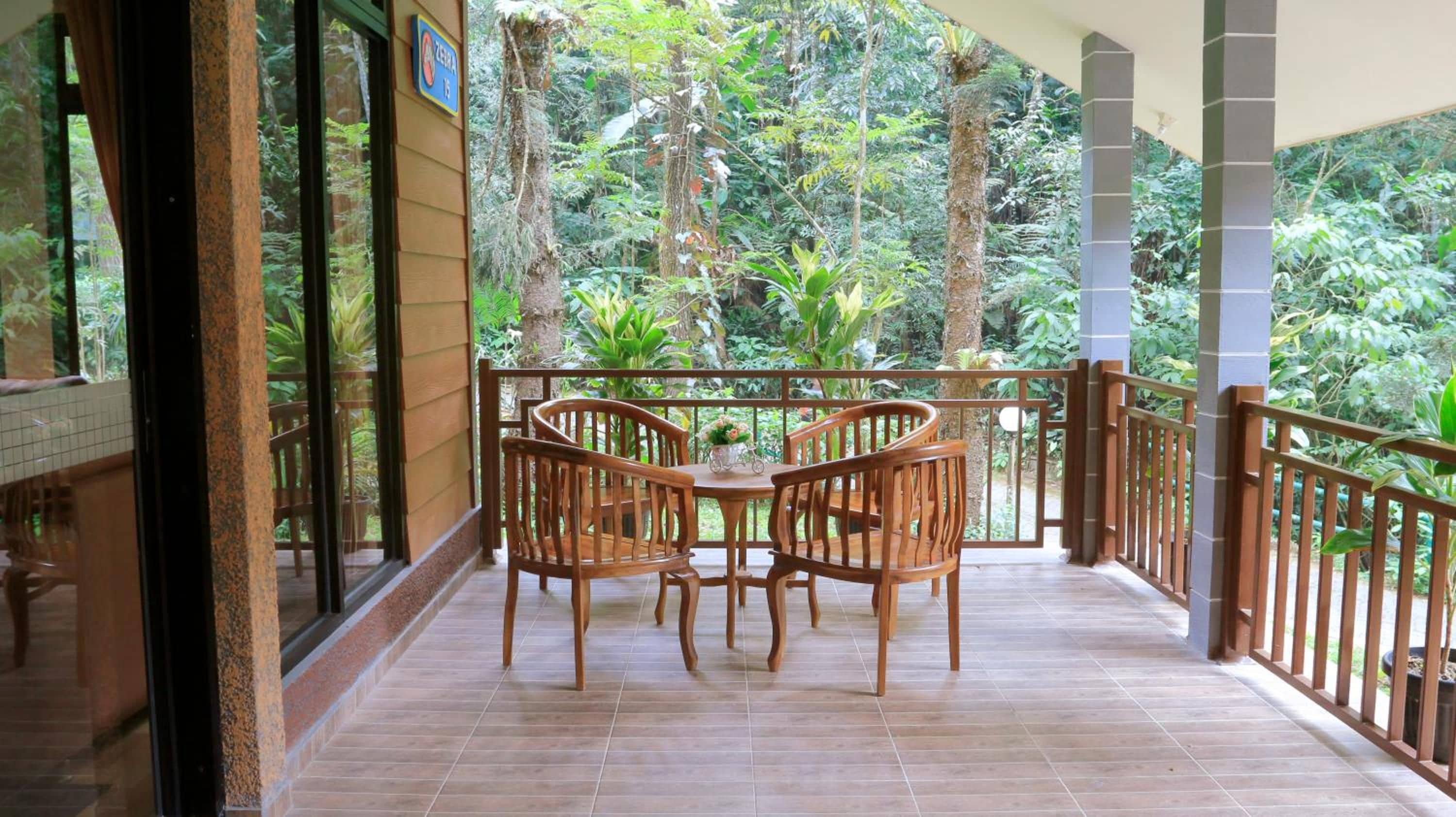 A balcony with wooden chairs and a table, surrounded by a railing, overlooking a lush garden.