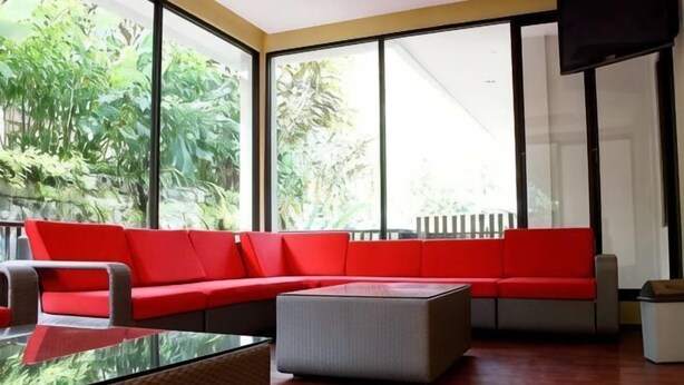 A modern living room with a red sectional sofa, a glass coffee table, and a grey ottoman. Large windows provide a view of lush greenery outside. - Safari Resort (Cisarua)
