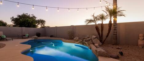 A swimming pool with a curved edge, surrounded by a patio area with chairs and a string of lights.