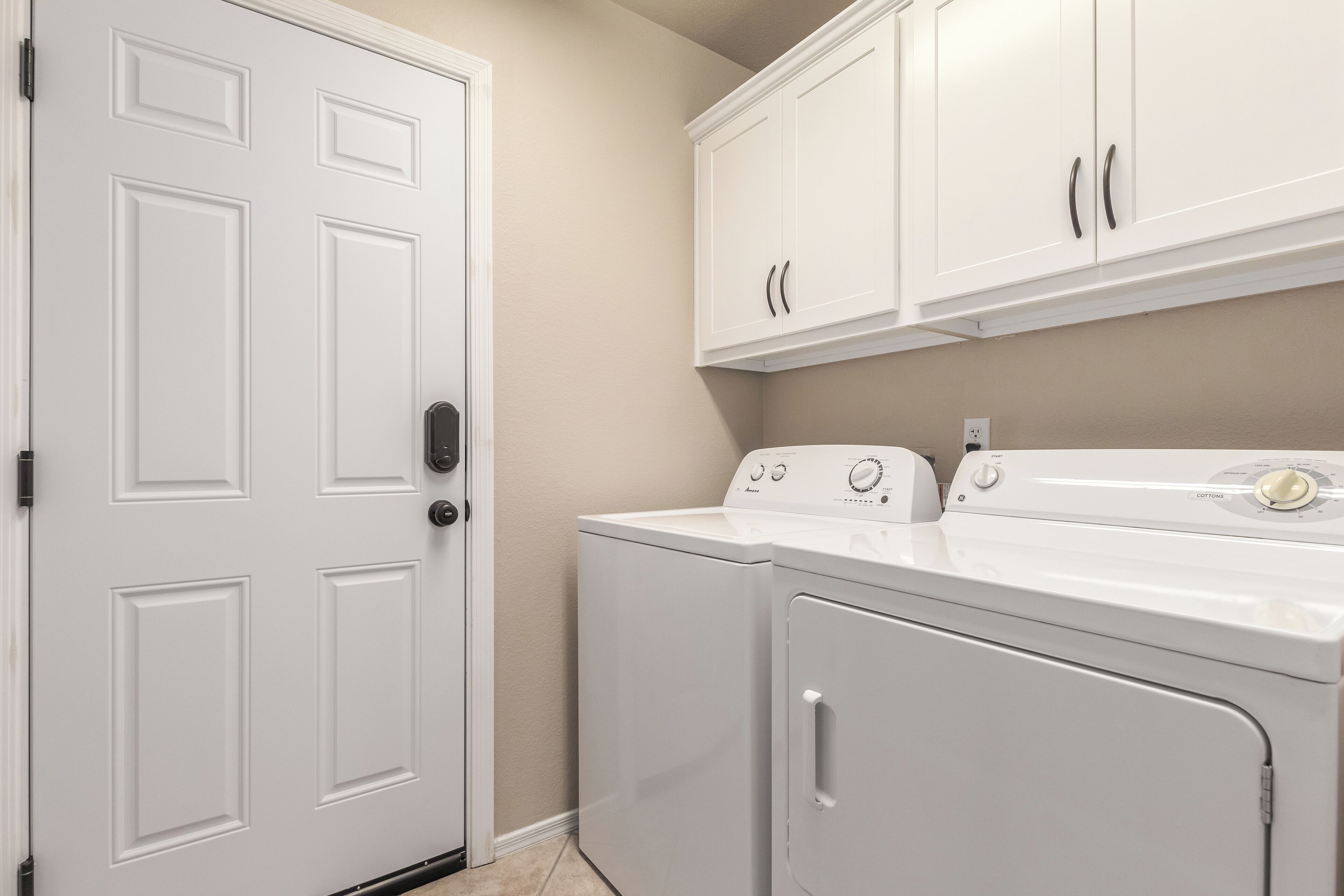 A laundry room with a white door, a washing machine, a dryer, and white cabinets above the appliances.
