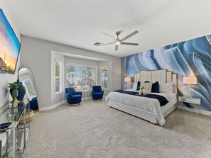A bedroom with a large bed, two armchairs, a ceiling fan, and a view of the outdoors through large windows. - Azurite Queen Creek (Queen Creek)