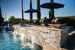Un espace aménagé au bord de la piscine, avec des chaises longues et des parasols, une cascade en pierre et un ciel dégagé.