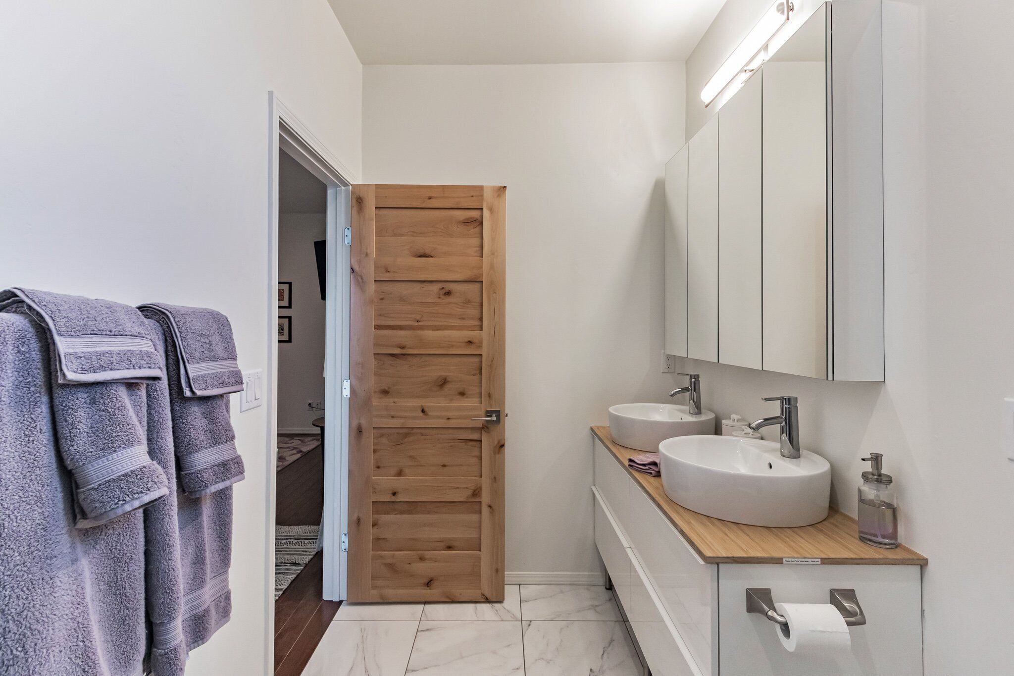 A modern bathroom with a wooden door, two sinks, a mirror, and towels on a rack.
