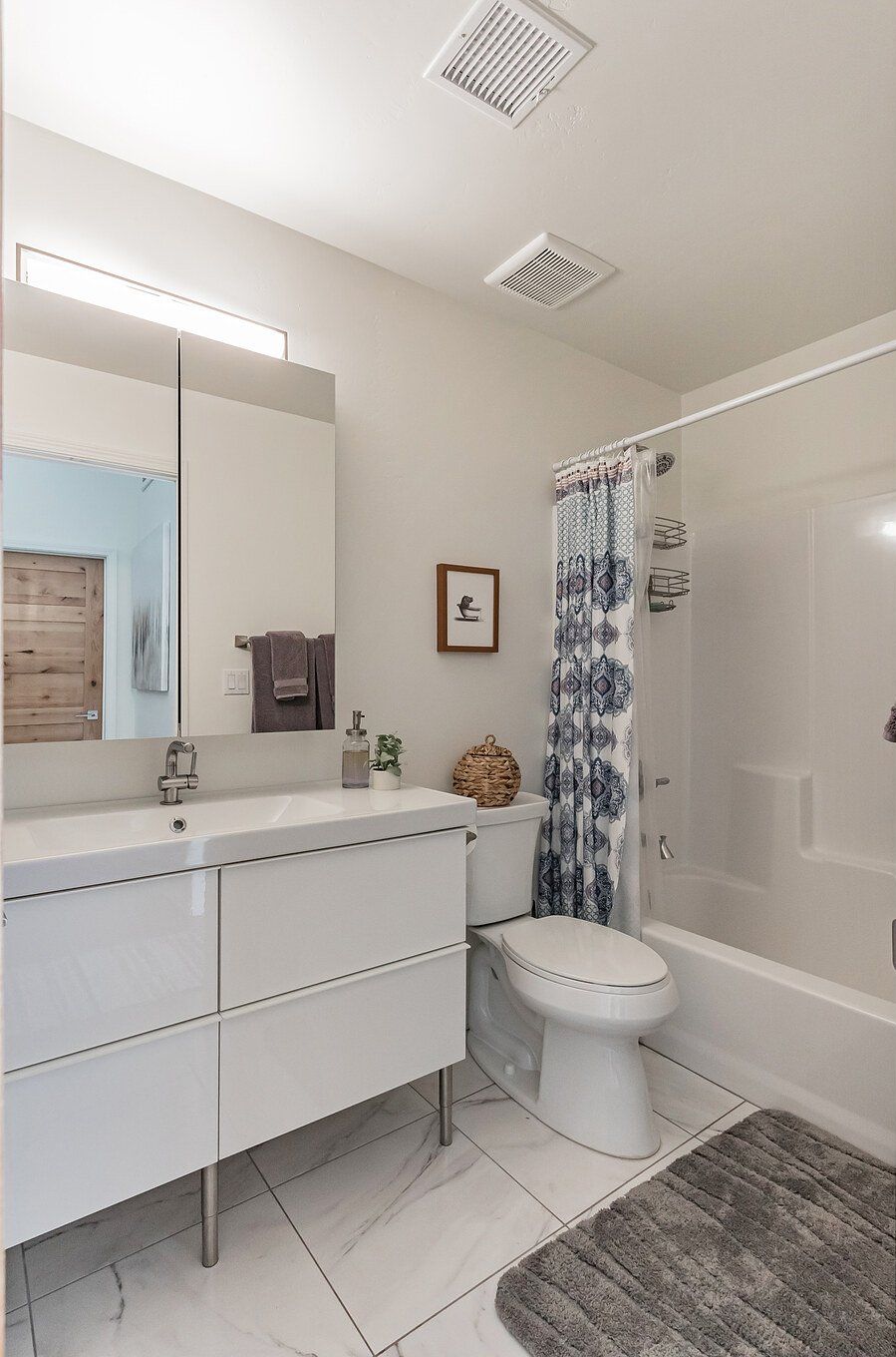 A modern bathroom with a white vanity, a toilet, a shower with a patterned curtain, and a mirror with a light above it.