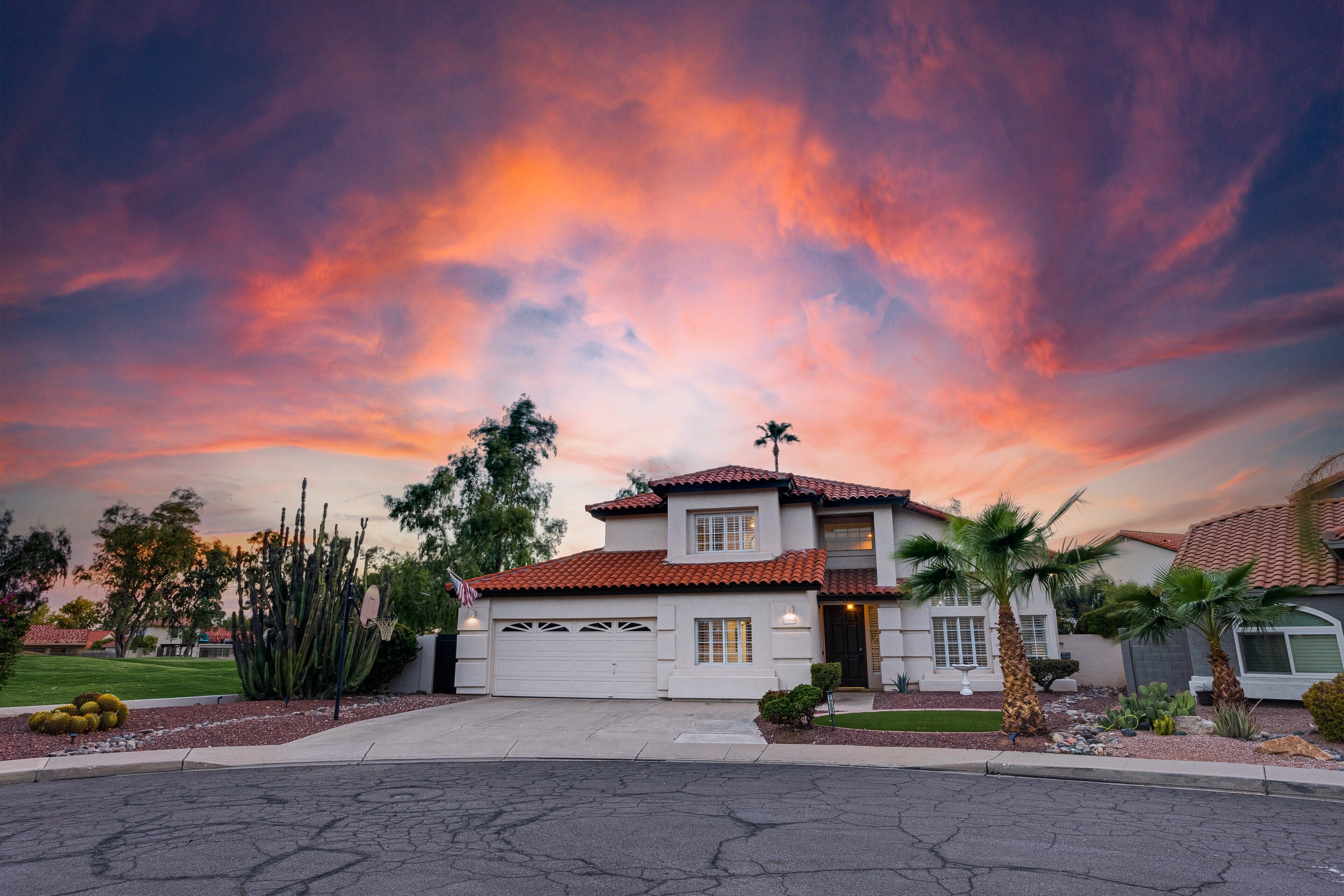 A two-story house with a red-tiled roof, a garage, and a front yard with cacti and palm trees.