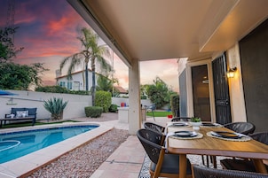 Um pátio com piscina, mesa de jantar e cadeiras, cercado por um jardim e uma casa.