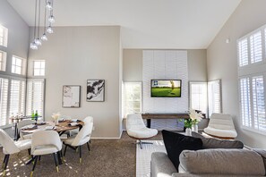 A modern living room with a dining area, a flat-screen TV mounted on the wall, and large windows with blinds. - 73rd Glendale (Glendale)