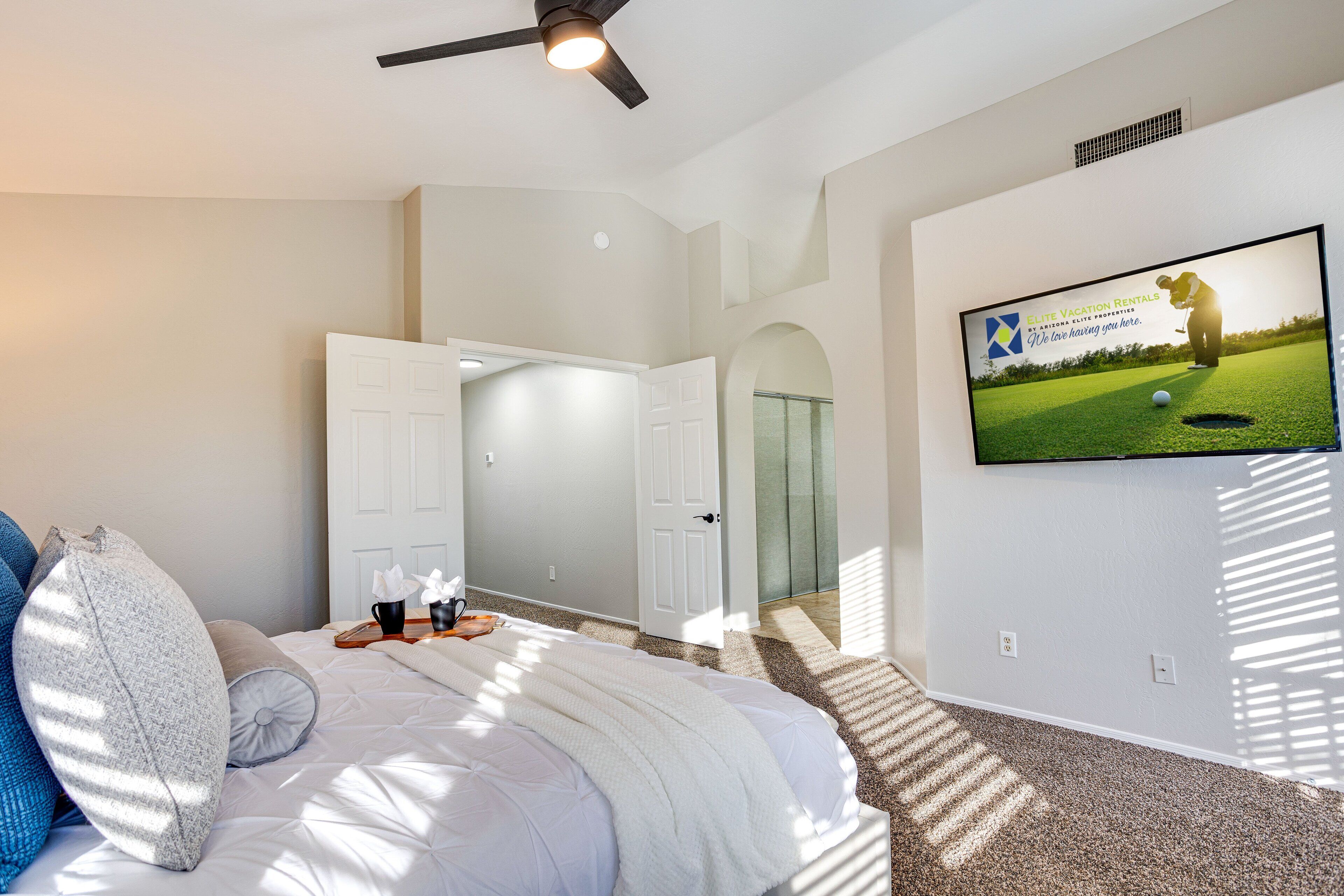 A bedroom with a bed, pillows, a TV on the wall, and a door leading to another room.