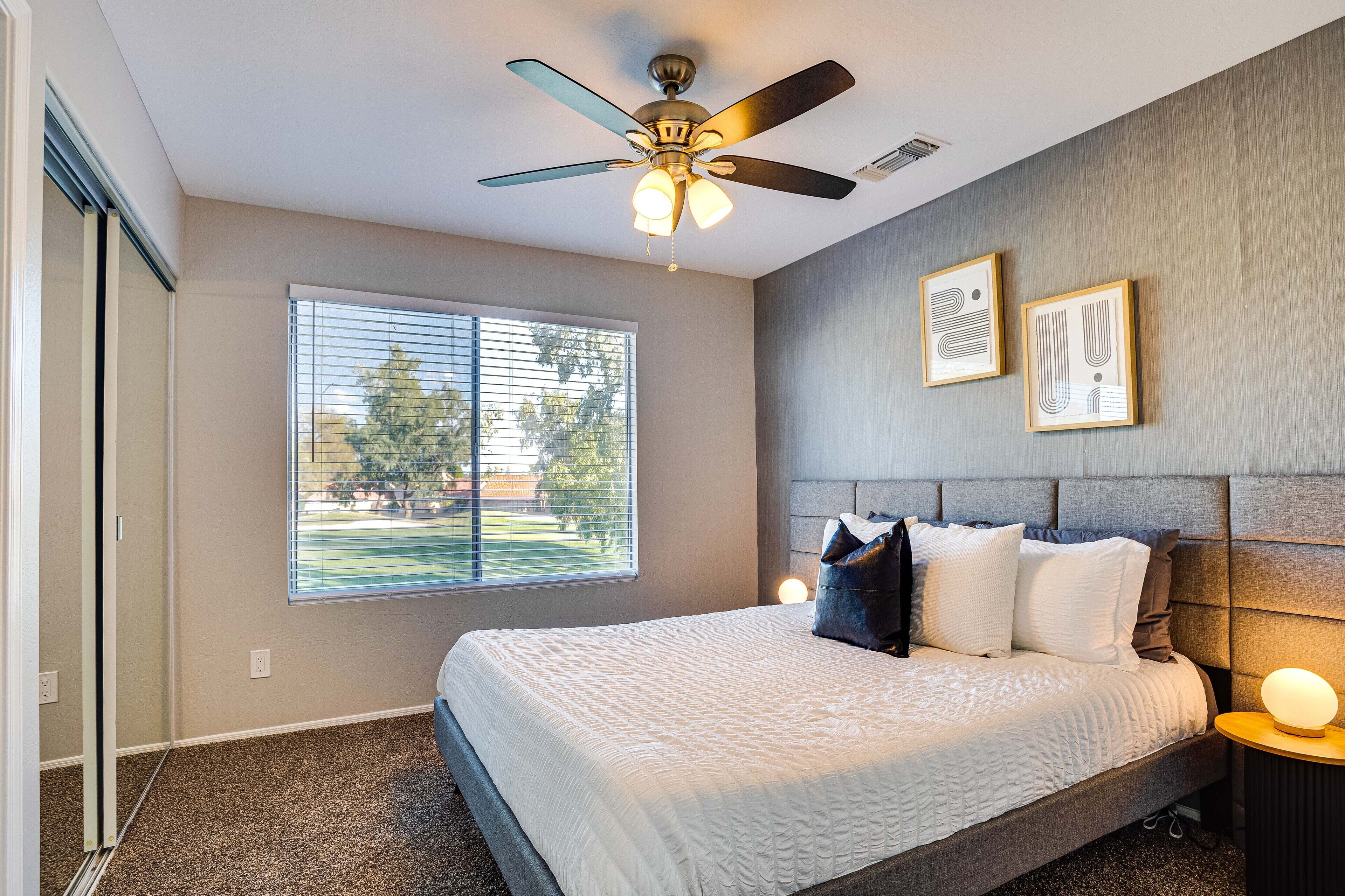 A bedroom with a bed, a ceiling fan, a window with blinds, and two framed pictures on the wall.