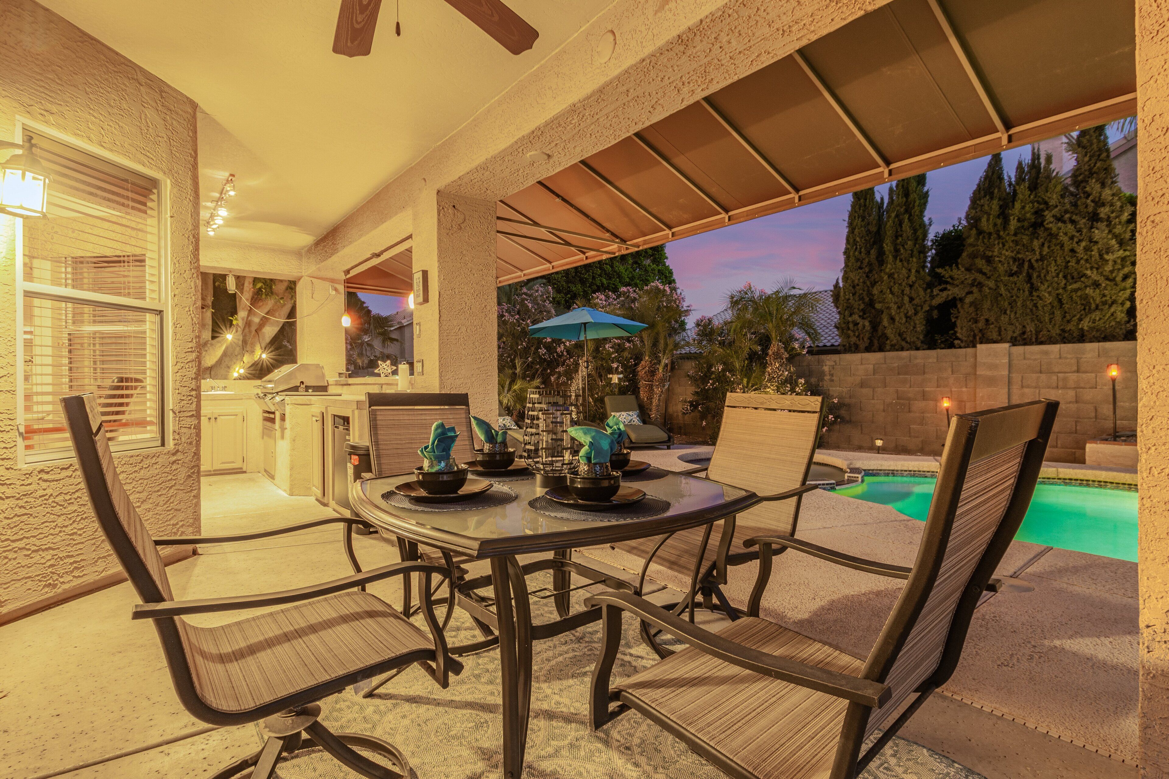Une terrasse extérieure avec une table et des chaises, une piscine et un espace abrité offrant une vue sur les arbres et le coucher du soleil.