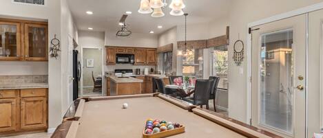 A pool table with a variety of balls, wooden cabinets, a kitchen area, and a dining space with chairs.