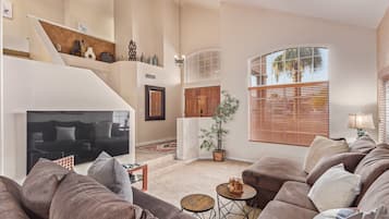 A spacious living room with a large sectional sofa, a flat-screen TV, a staircase, and a large window with blinds.