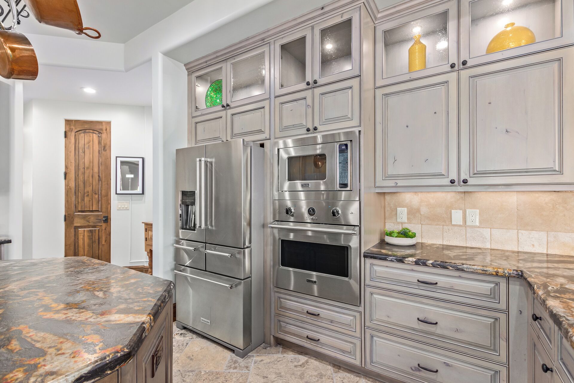 A kitchen with a stainless steel refrigerator, double oven, and a microwave. There are cabinets with glass doors and a marble countertop.