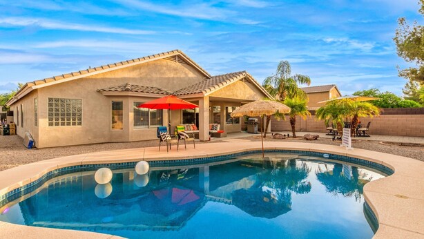 Una casa con piscina, mobiliario de patio y un paraguas de paja.