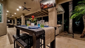 An outdoor dining area with a table set for two, surrounded by wicker chairs, a ceiling fan, and a TV mounted on a pillar displaying a scenic view. - Northridge Mesa (Mesa)