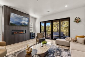 A modern living room with a fireplace, comfortable seating, and a large window with a view of trees. - Cottonrose Flagstaff (Flagstaff)