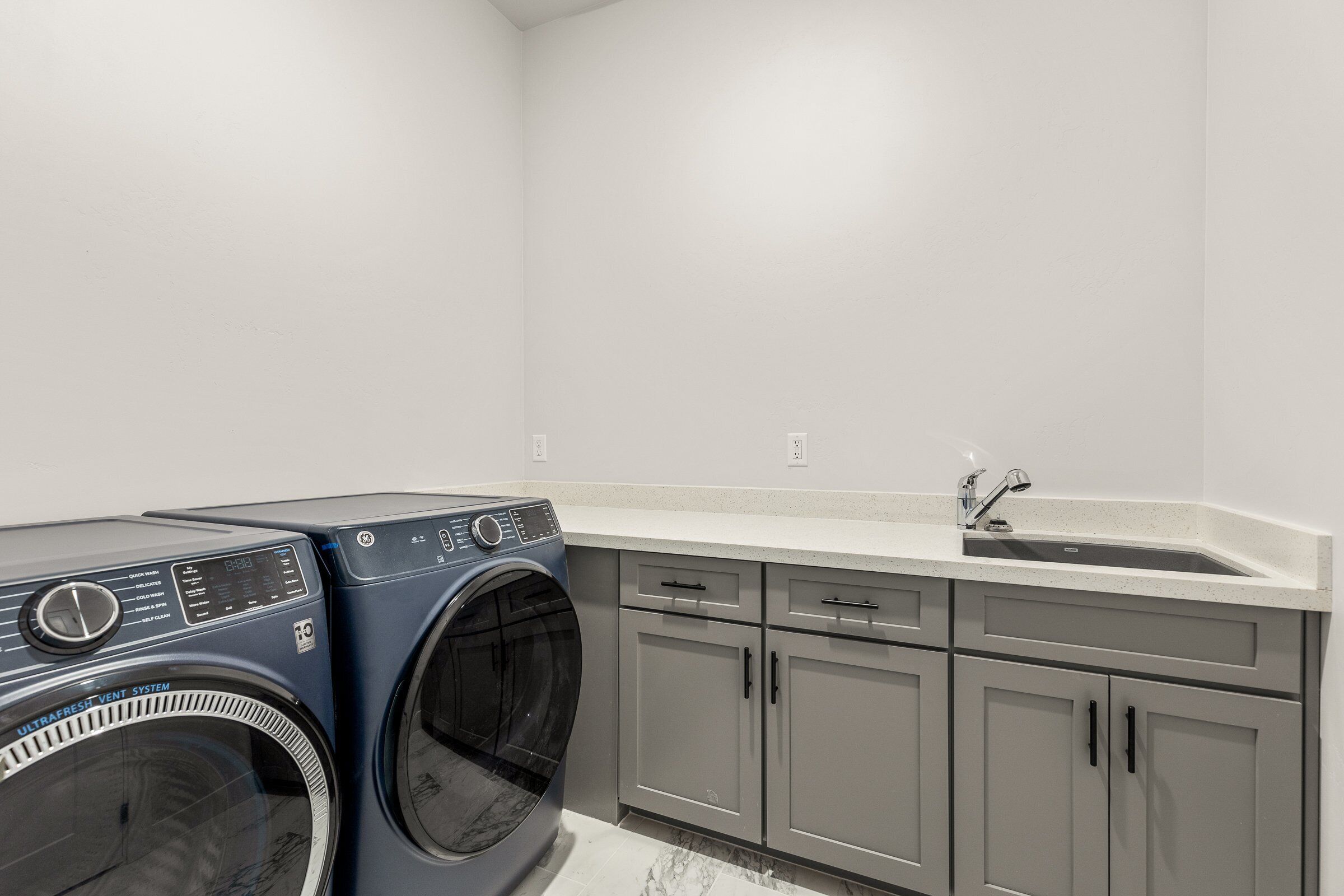 A laundry area with a front-loading washer and dryer, a sink, and grey cabinets.