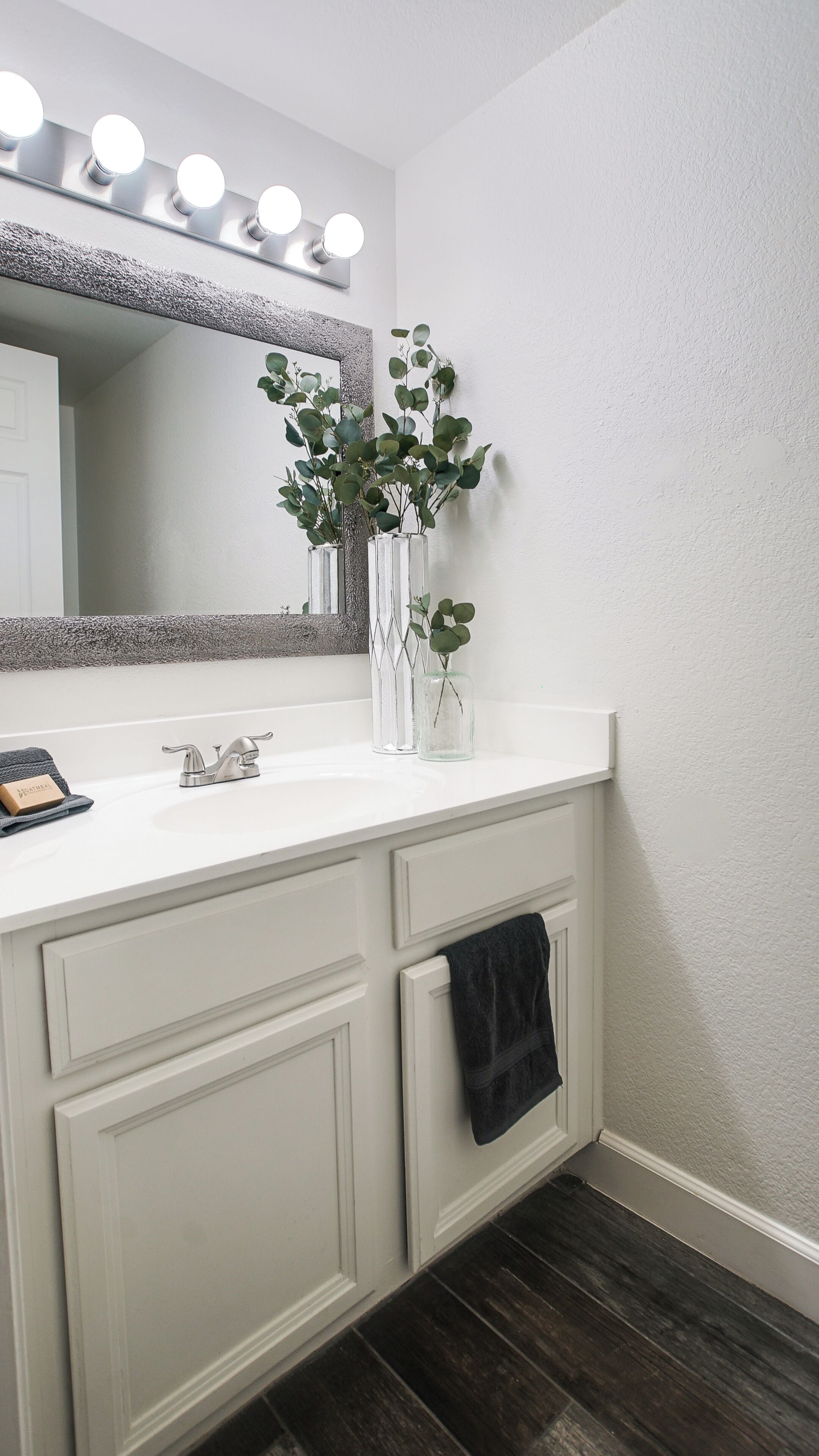 A bathroom with a white vanity, a mirror with lights, and a plant in a glass vase.
