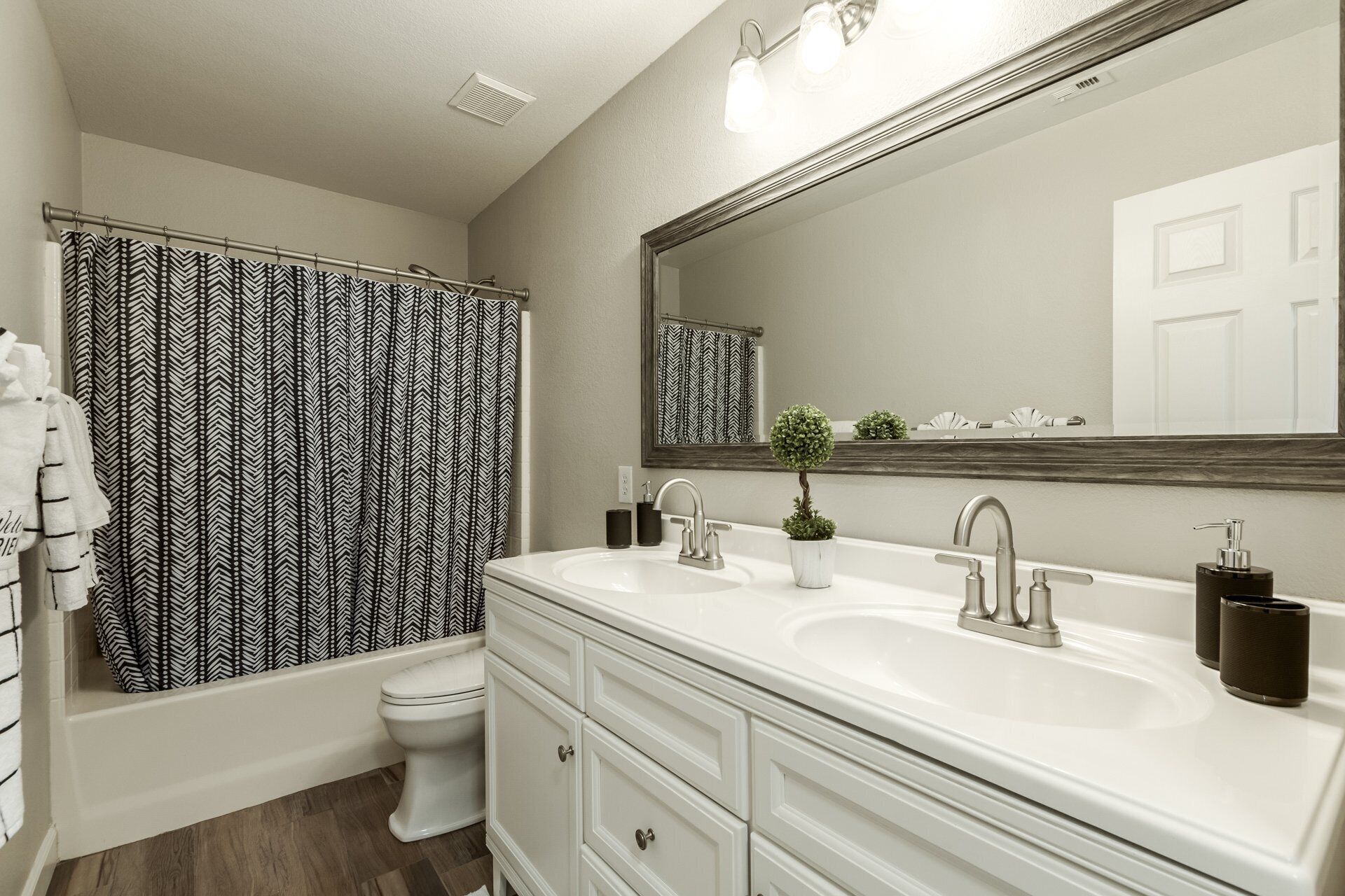 Une salle de bain avec un meuble-lavabo blanc, deux vasques, un miroir, un rideau de douche à motifs noir et blanc et des toilettes.