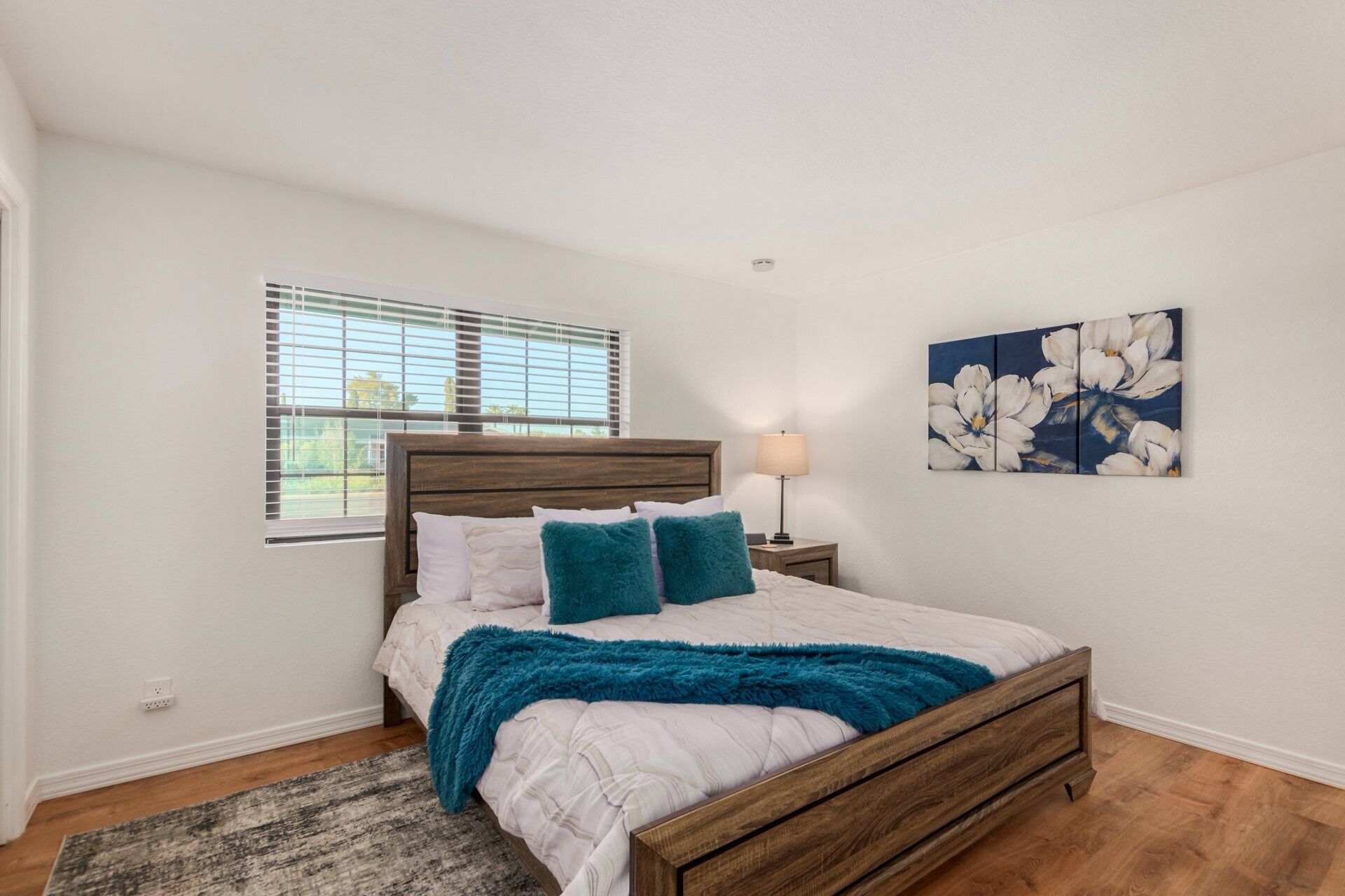 A bedroom with a wooden bed, white bedding, teal pillows, a blue throw blanket, a nightstand with a lamp, and a floral painting on the wall.