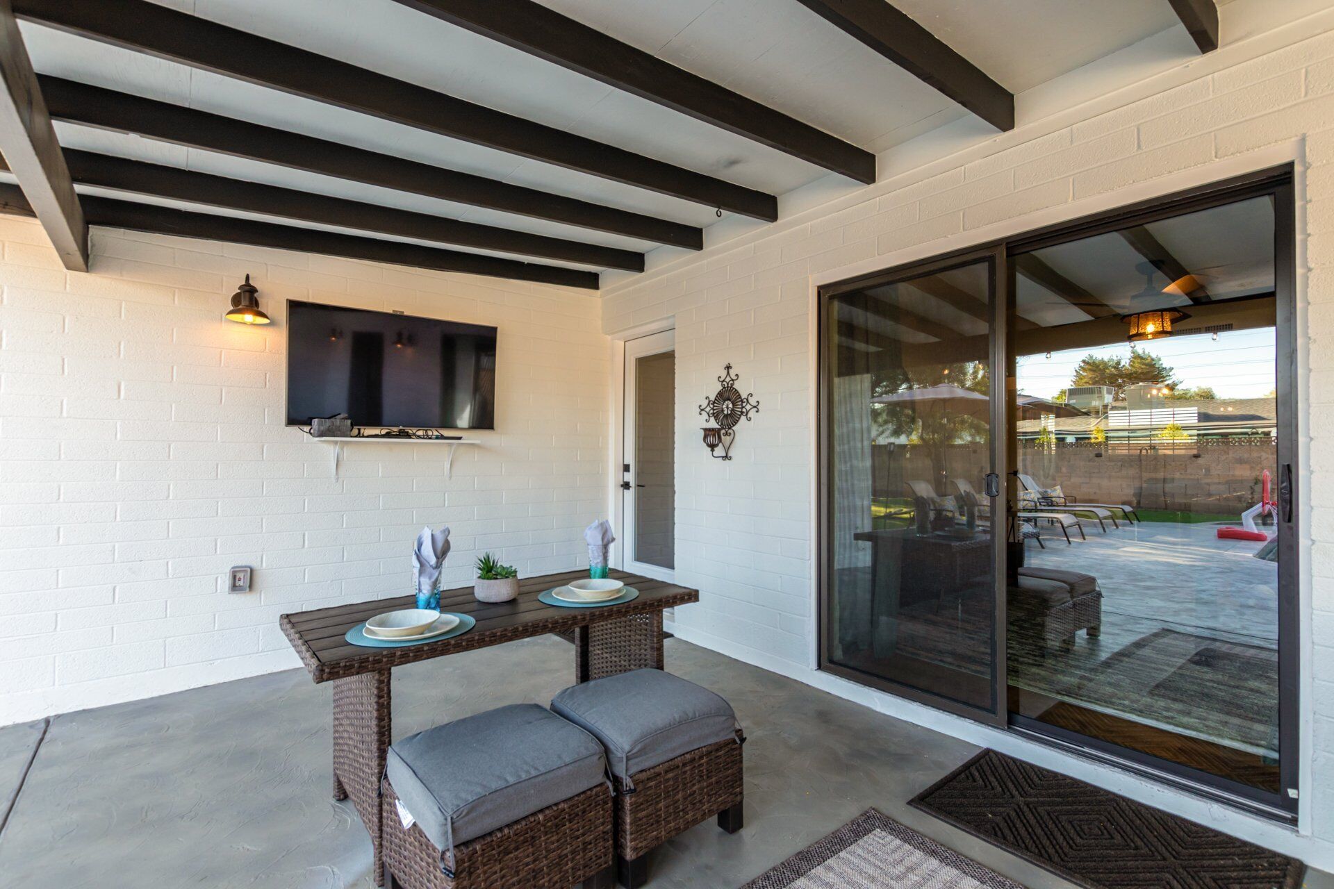 Un patio couvert comprenant une table et deux fauteuils rembourrés, un téléviseur fixé au mur et une grande porte-fenêtre coulissante donnant sur un espace extérieur aménagé pour s’asseoir.