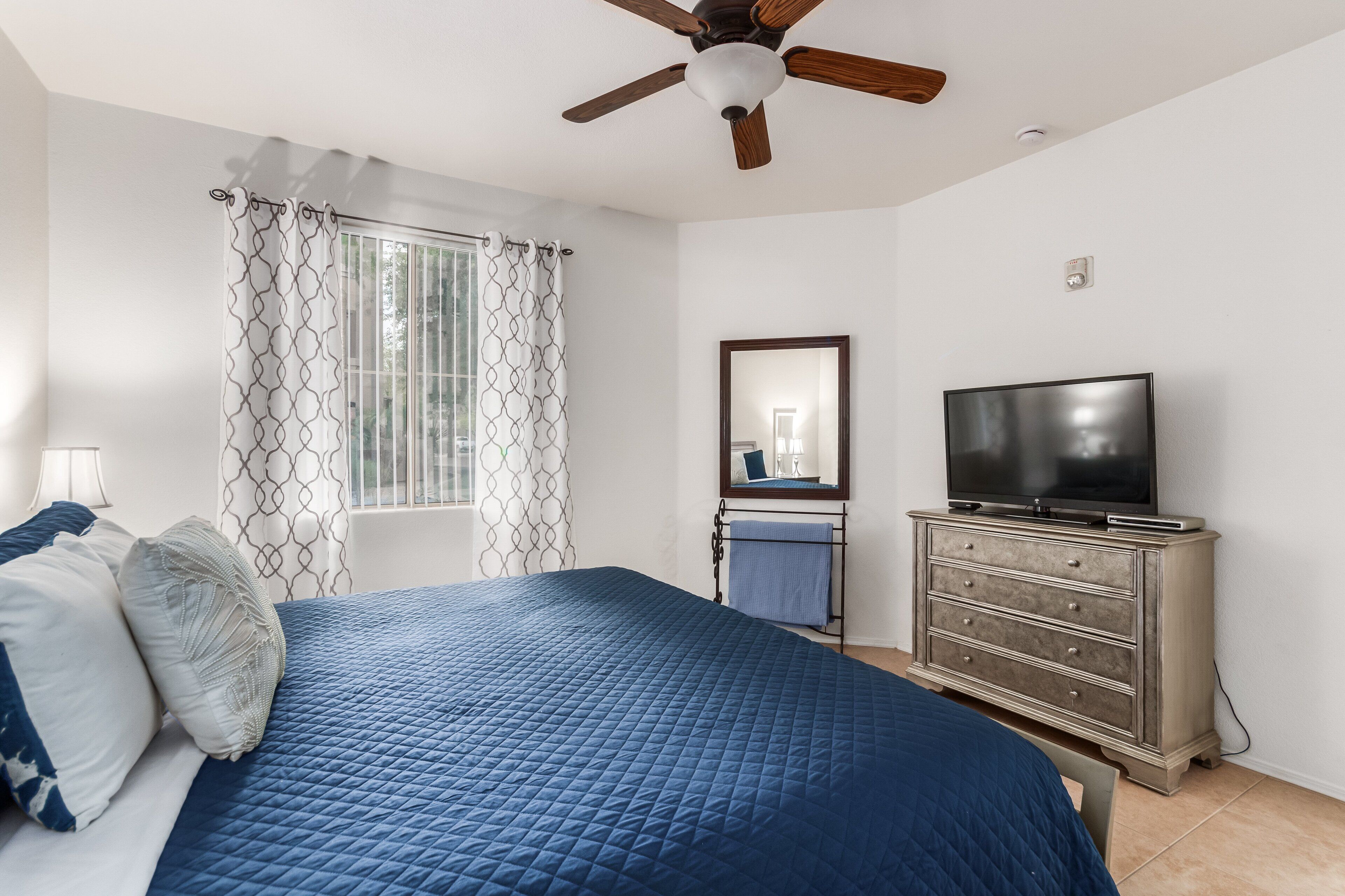 Une chambre à coucher comprenant un lit, une commode, un téléviseur, un ventilateur de plafond et une fenêtre avec des rideaux.