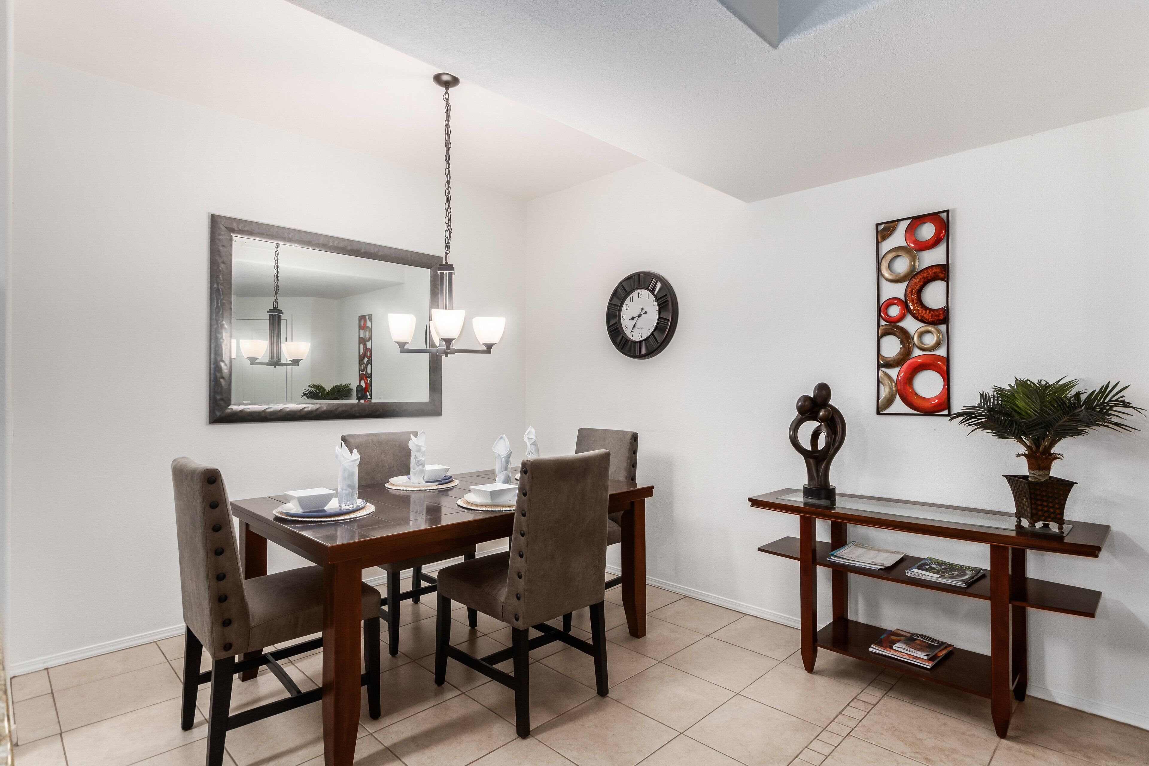 A dining area with a wooden table, four chairs, a chandelier, a wall clock, and a decorative art piece.