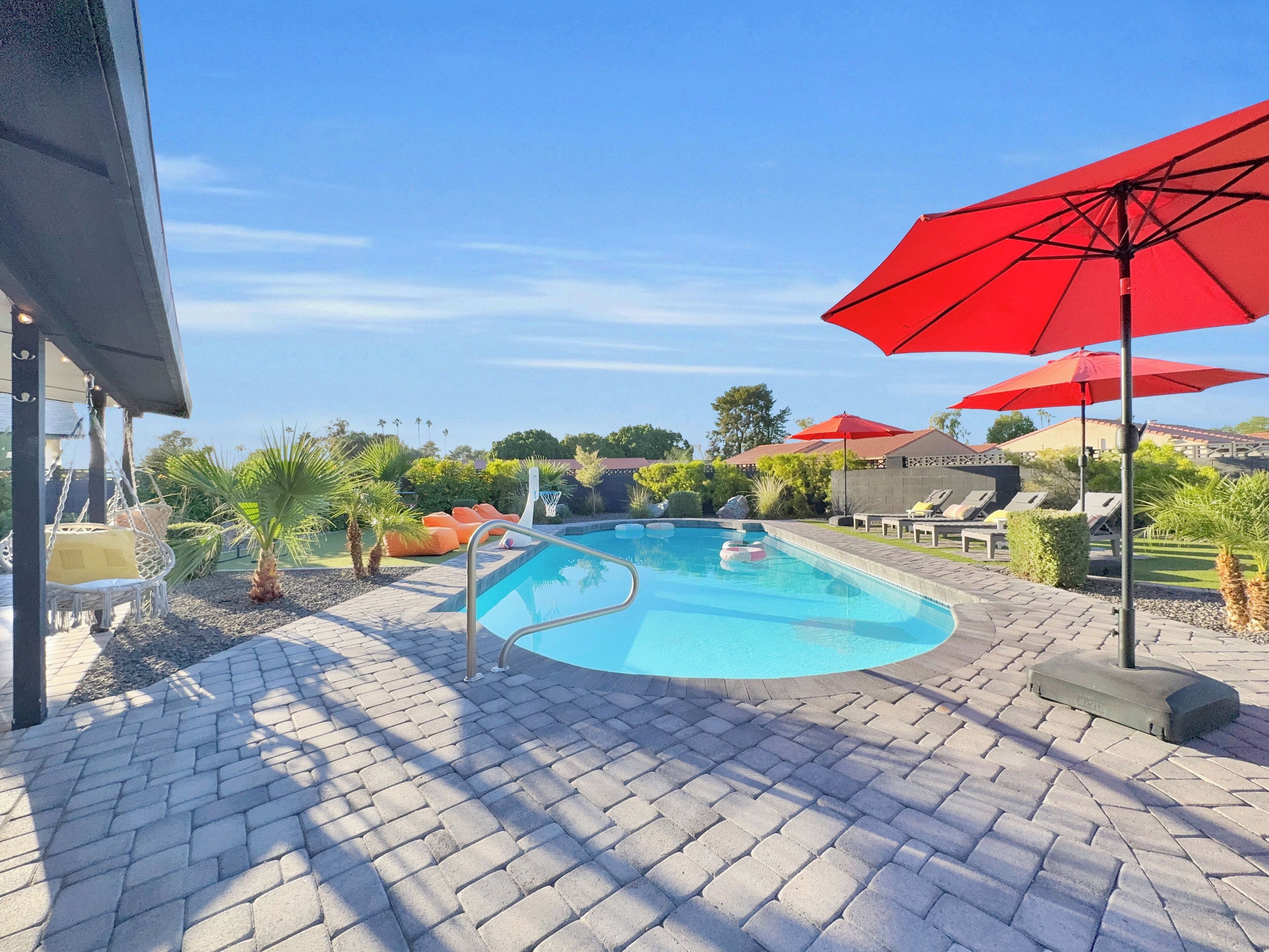 Uma piscina com um terraço de tijolos, espreguiçadeiras e guarda-sóis vermelhos.