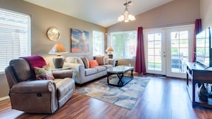 A living room with a sofa, coffee table, and lamps. There is a clock on the wall and a painting above the sofa.