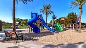 A playground with a blue slide and a green slide, surrounded by palm trees and a bench.