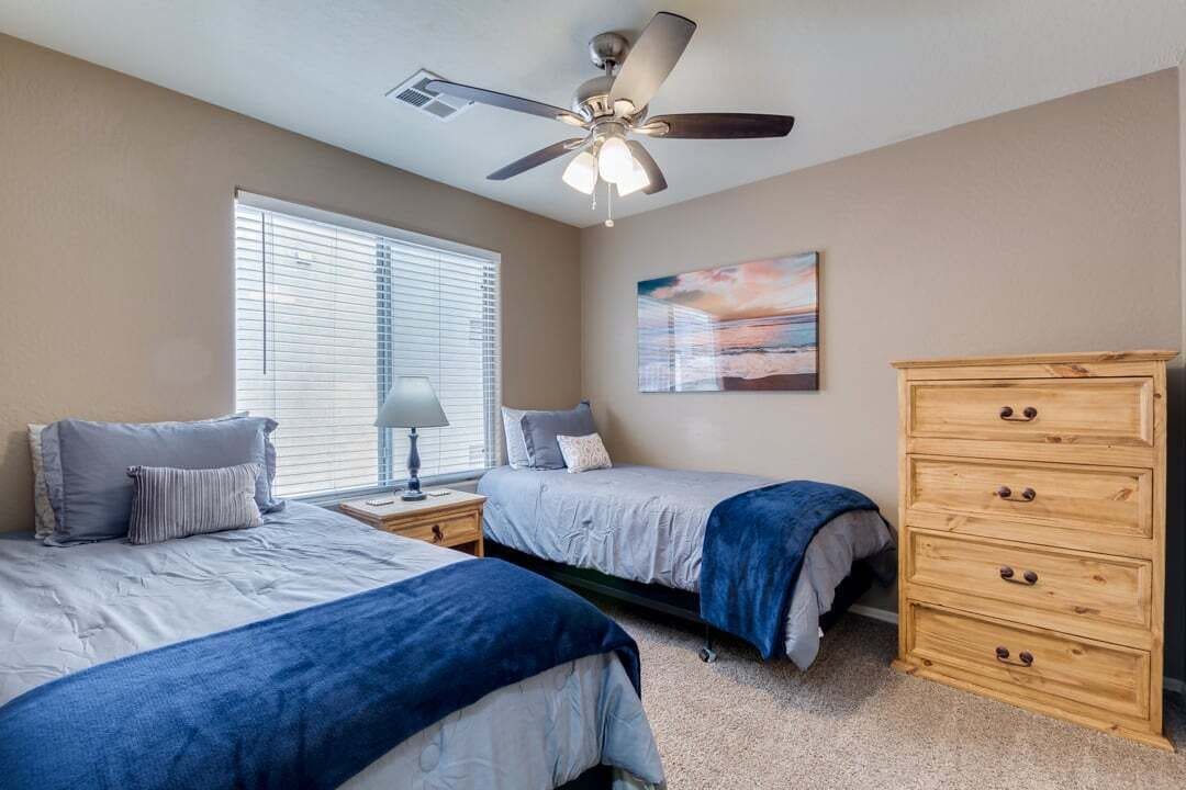 A bedroom with two beds, a ceiling fan, a nightstand with a lamp, a dresser, and a window with blinds.