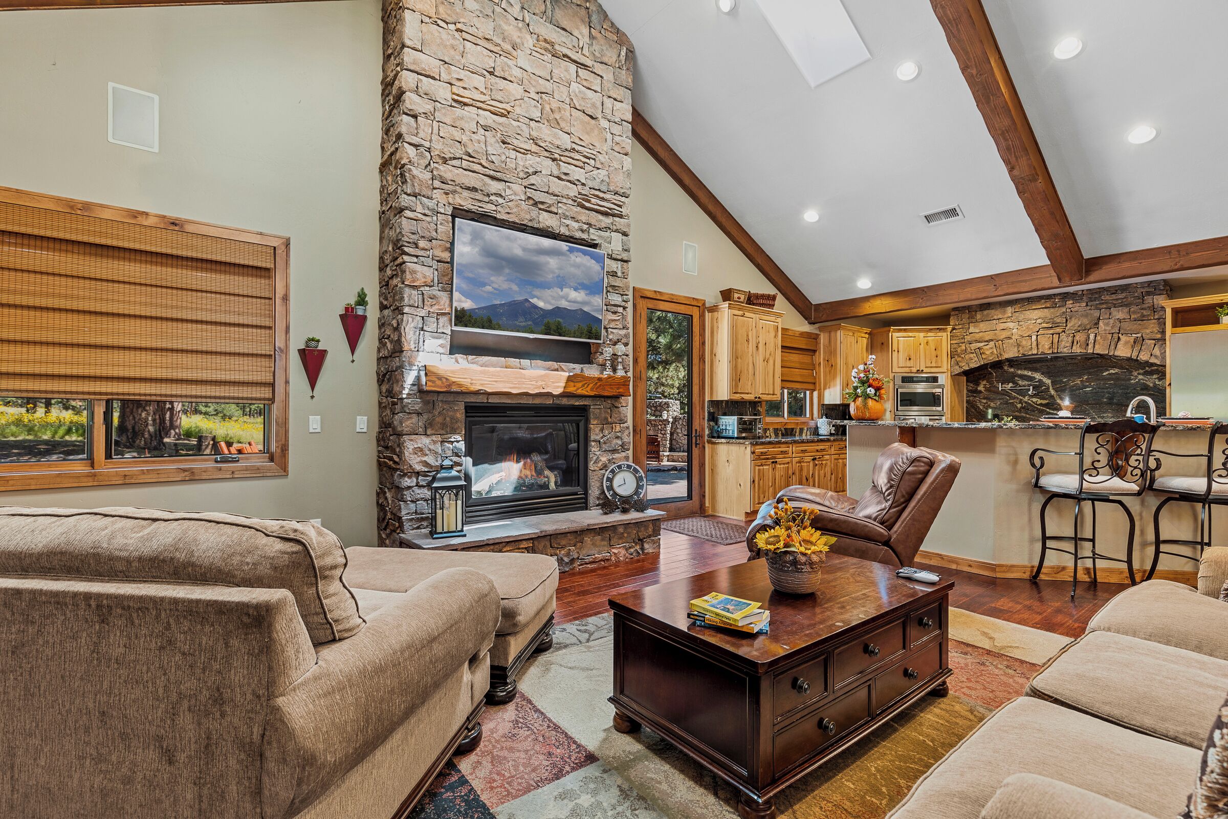 A cozy living room with a stone fireplace, wooden furniture, and a kitchen area with a stone backsplash.