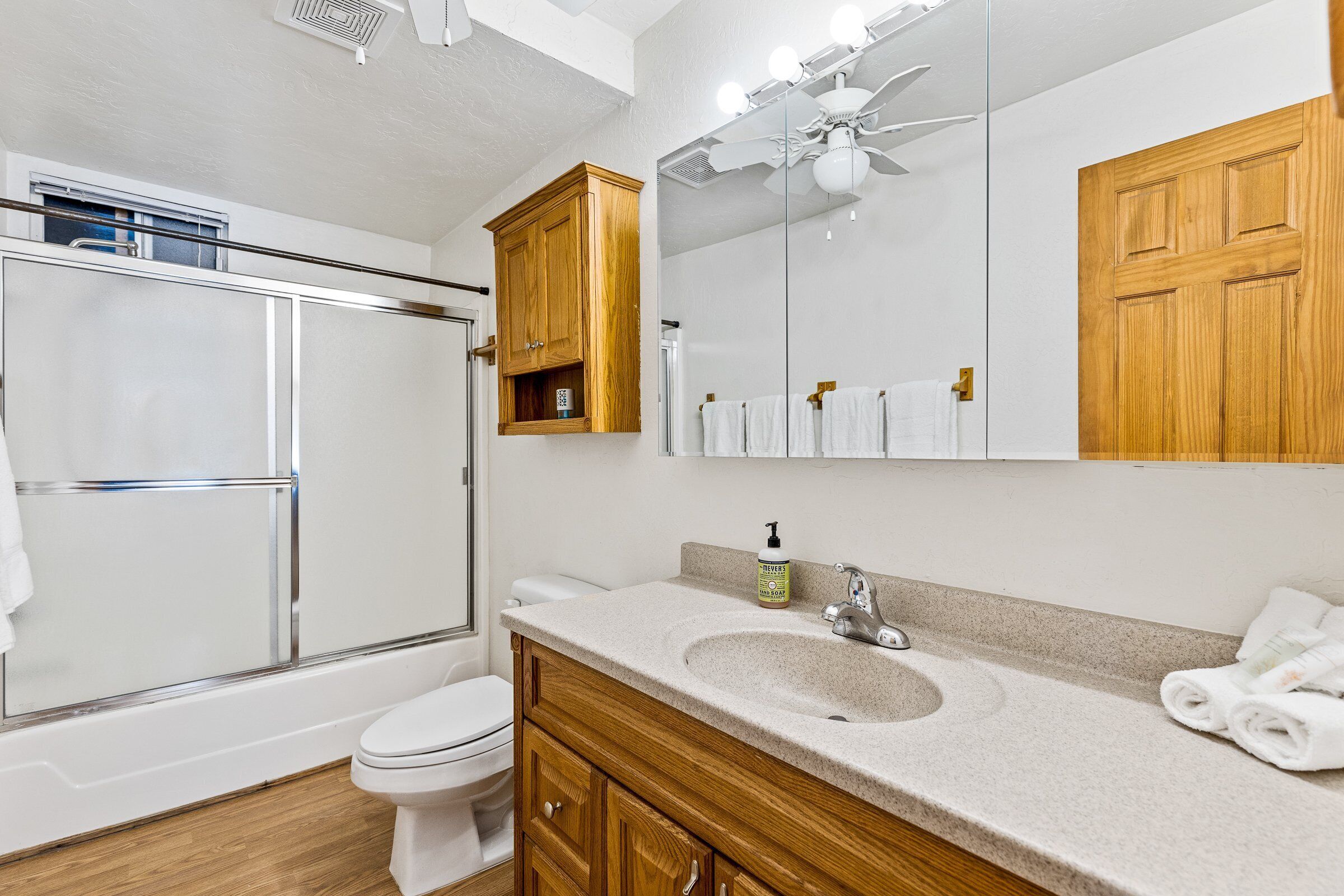 Un baño moderno con mampara de ducha de vidrio, inodoro blanco, armarios de madera, un espejo grande y un ventilador de techo.