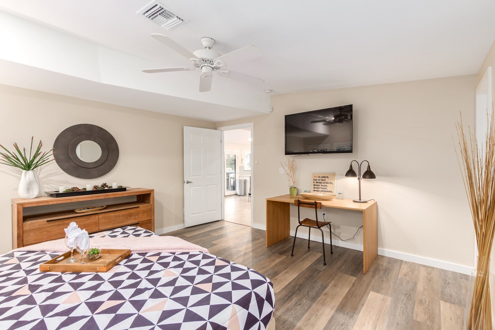 A modern bedroom with a geometric-patterned bedspread, a wooden nightstand, a desk with a chair, a television, and a ceiling fan.
