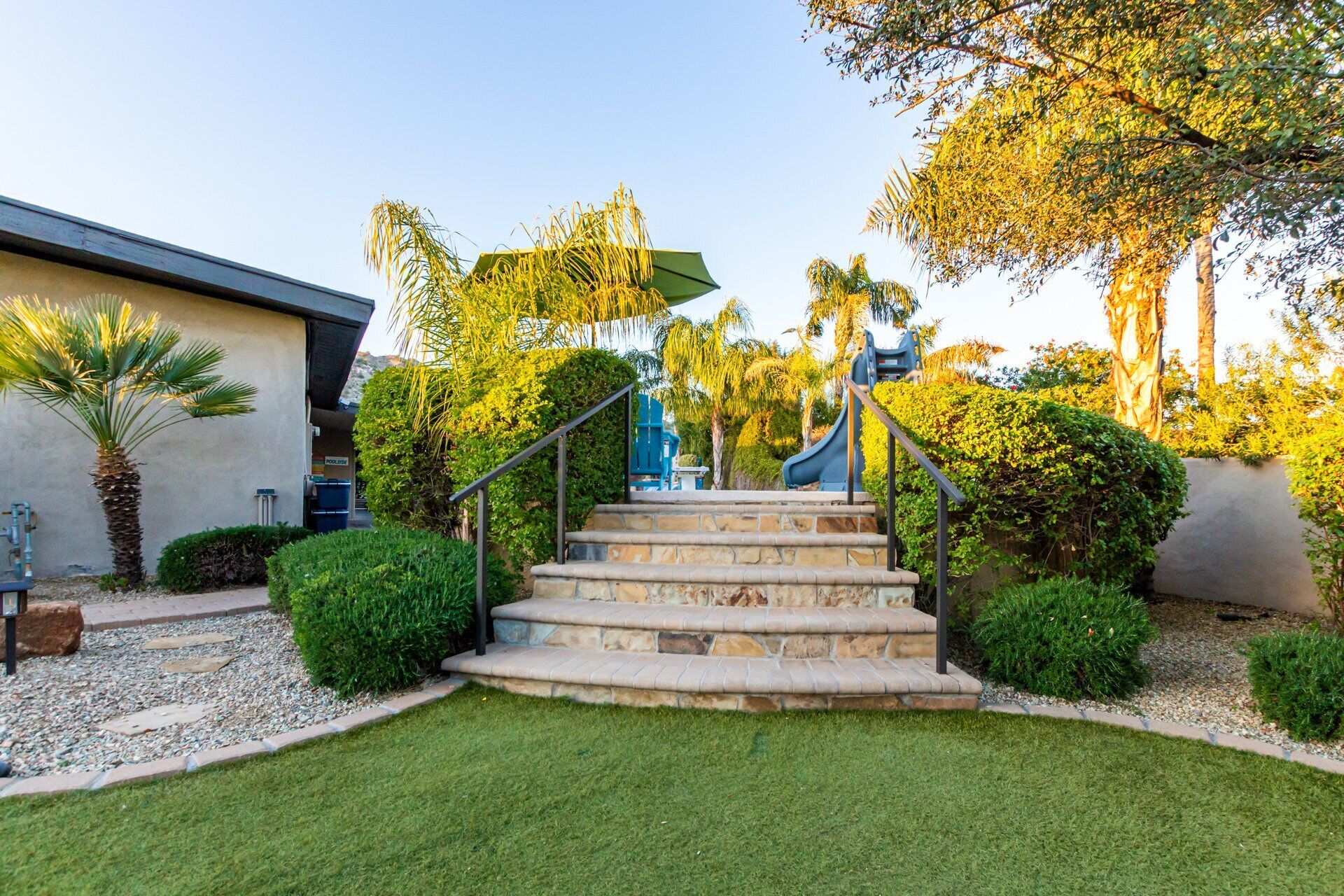 A set of stone steps leading up to a blue slide, surrounded by well-maintained grass and various shrubs.