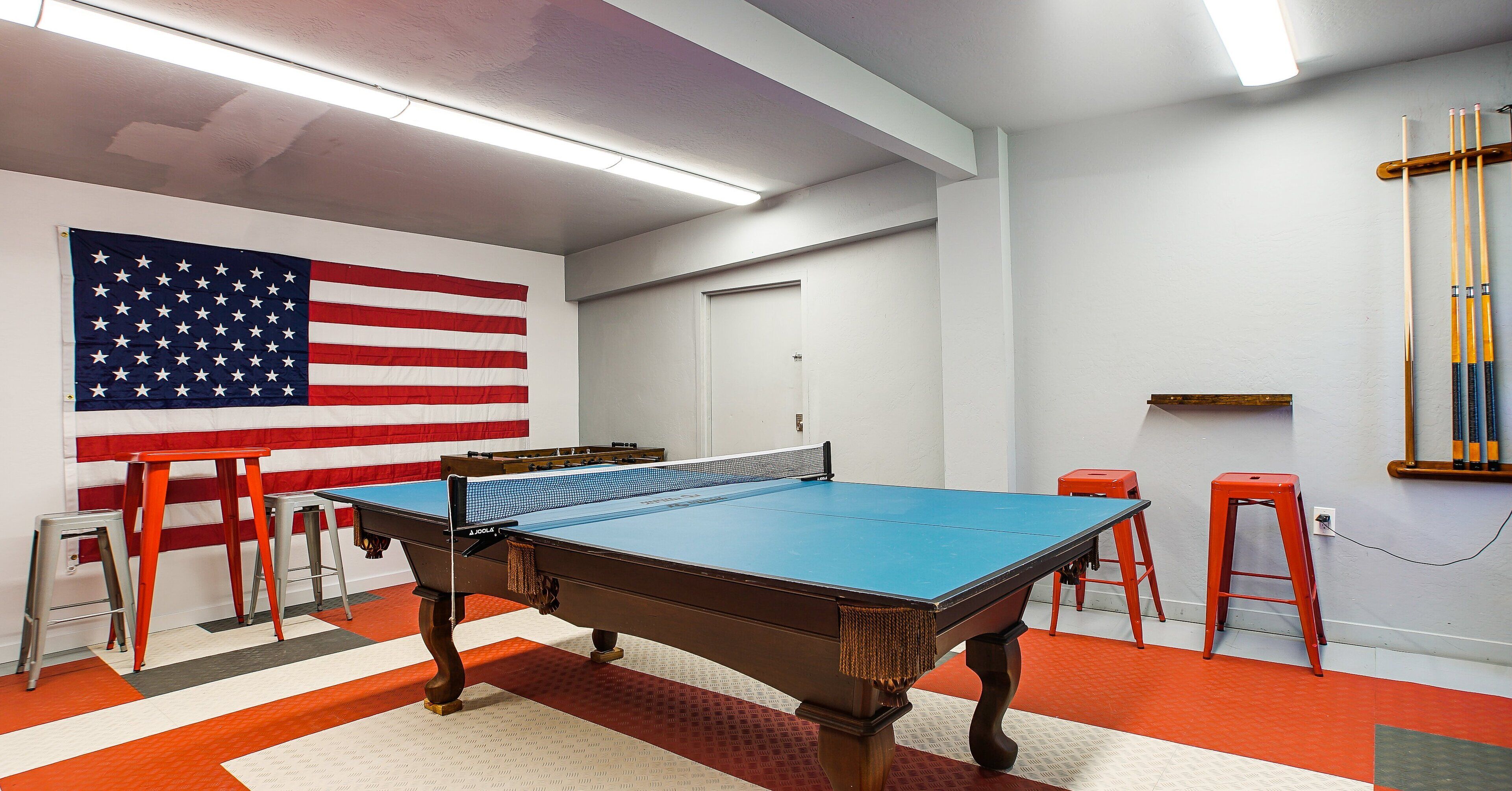 A pool table with a blue felt surface, surrounded by red stools, in a room with a large American flag on the wall.