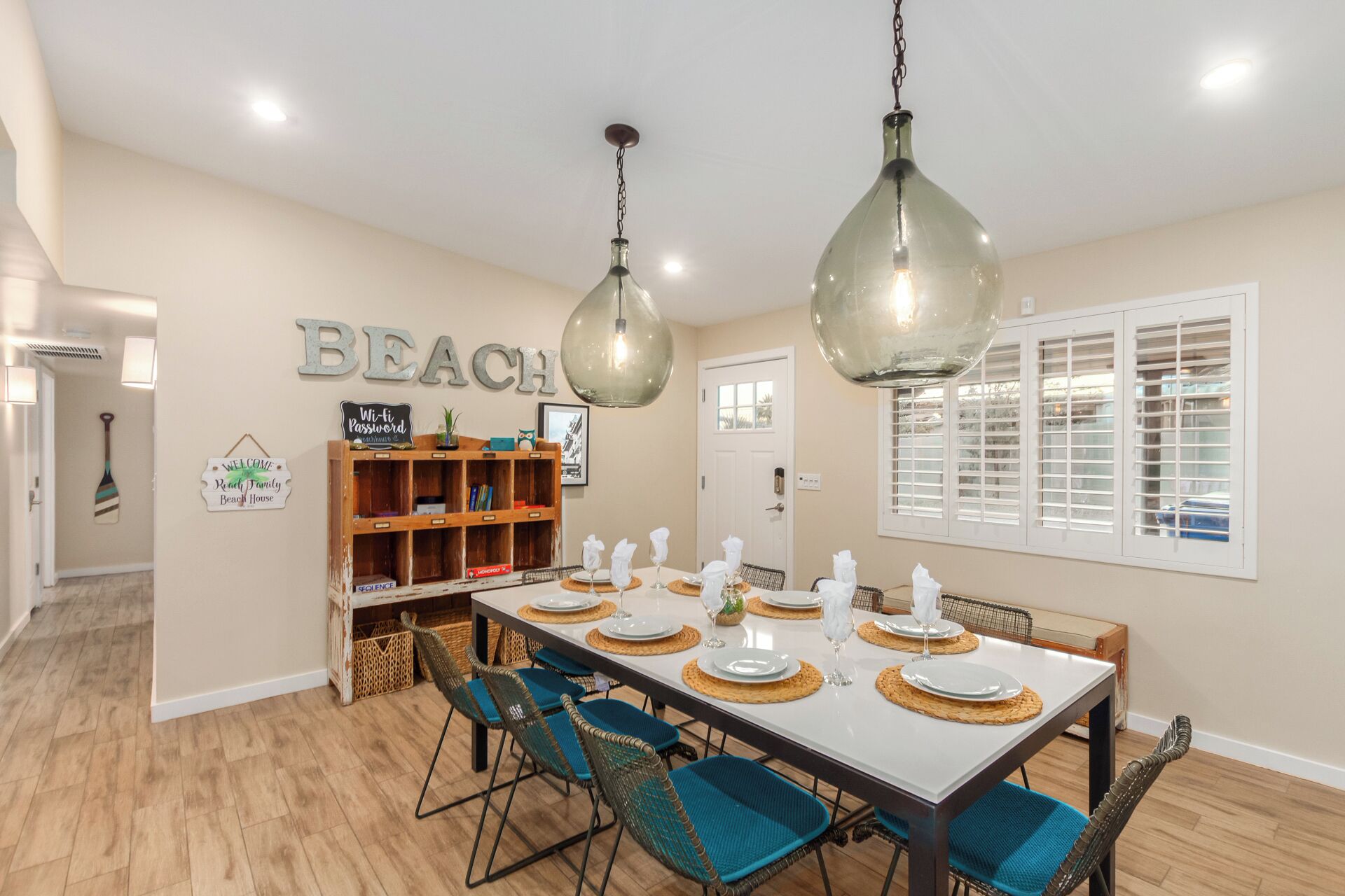 Un coin repas avec une longue table dressée pour six personnes, entourée de chaises en osier avec des coussins bleus. Une étagère en bois, sur laquelle sont disposés divers objets, et une enseigne indiquant « PLAGE » sont accrochées au mur.