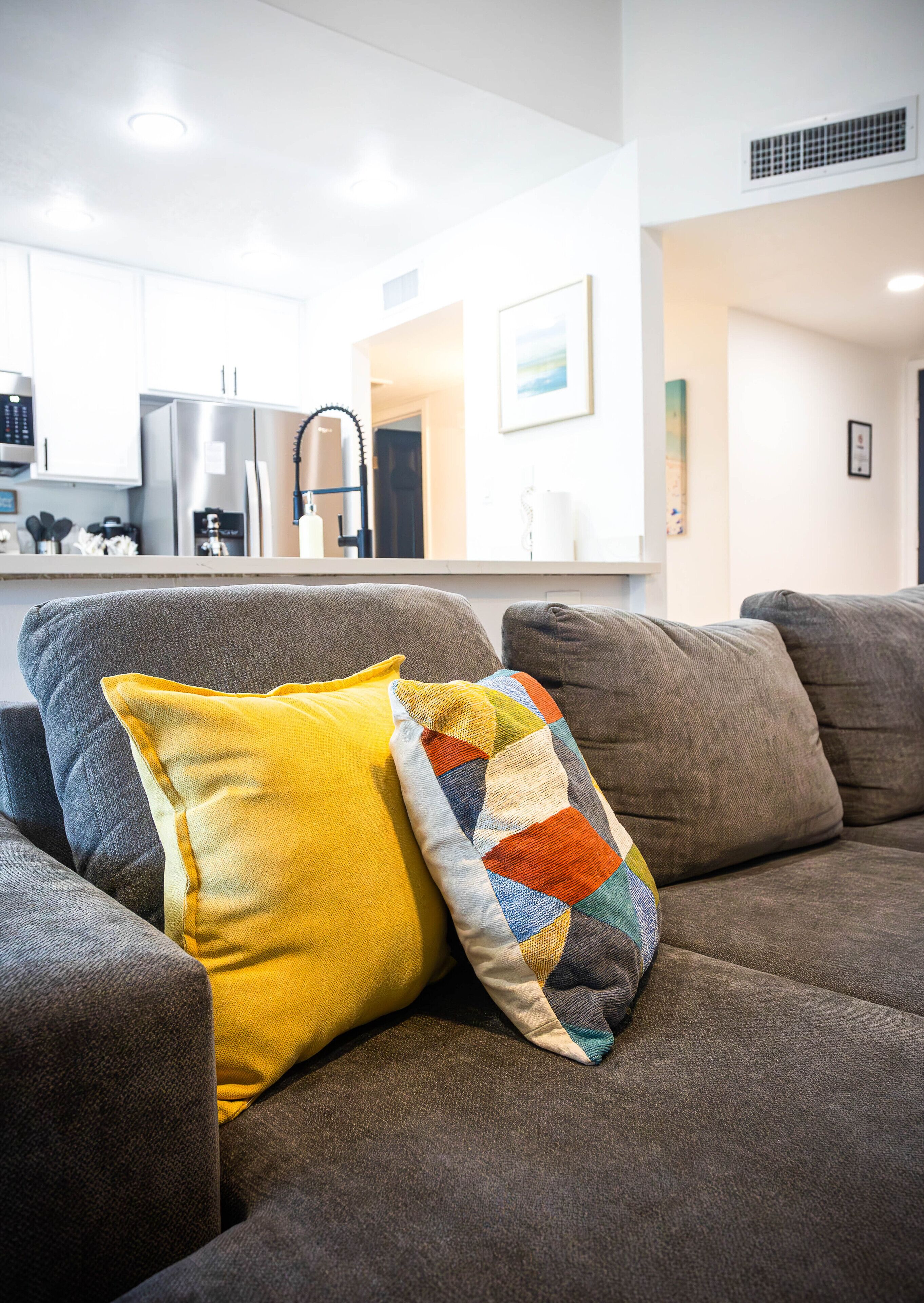 Un salon moderne avec un canapé gris, un coussin jaune et multicolore, et une cuisine en arrière-plan.