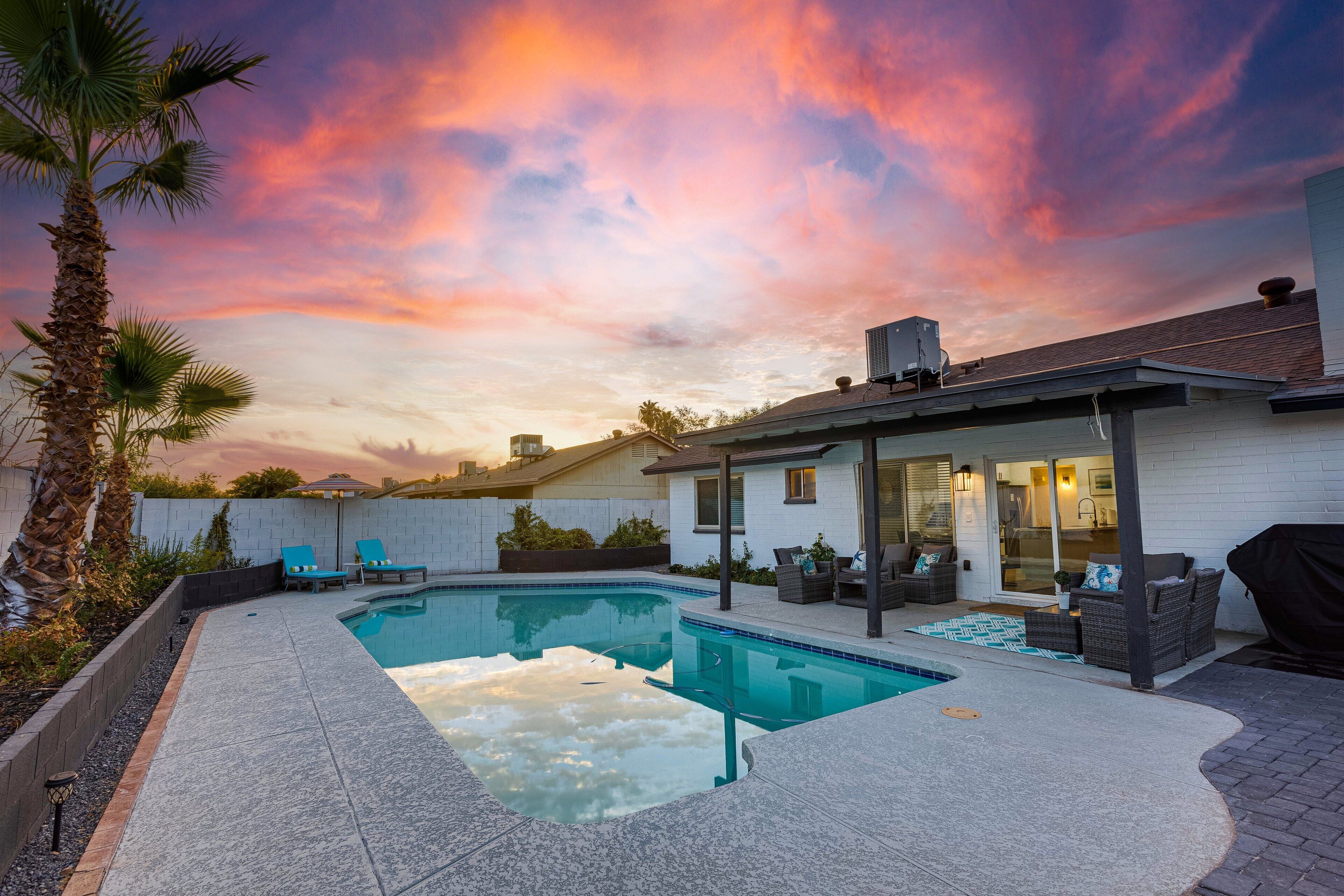 A swimming pool with lounge chairs, a covered patio, and a view of the sunset.