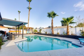 En swimmingpool med en pergola och solstolar, omgiven av palmer och en vit vägg.