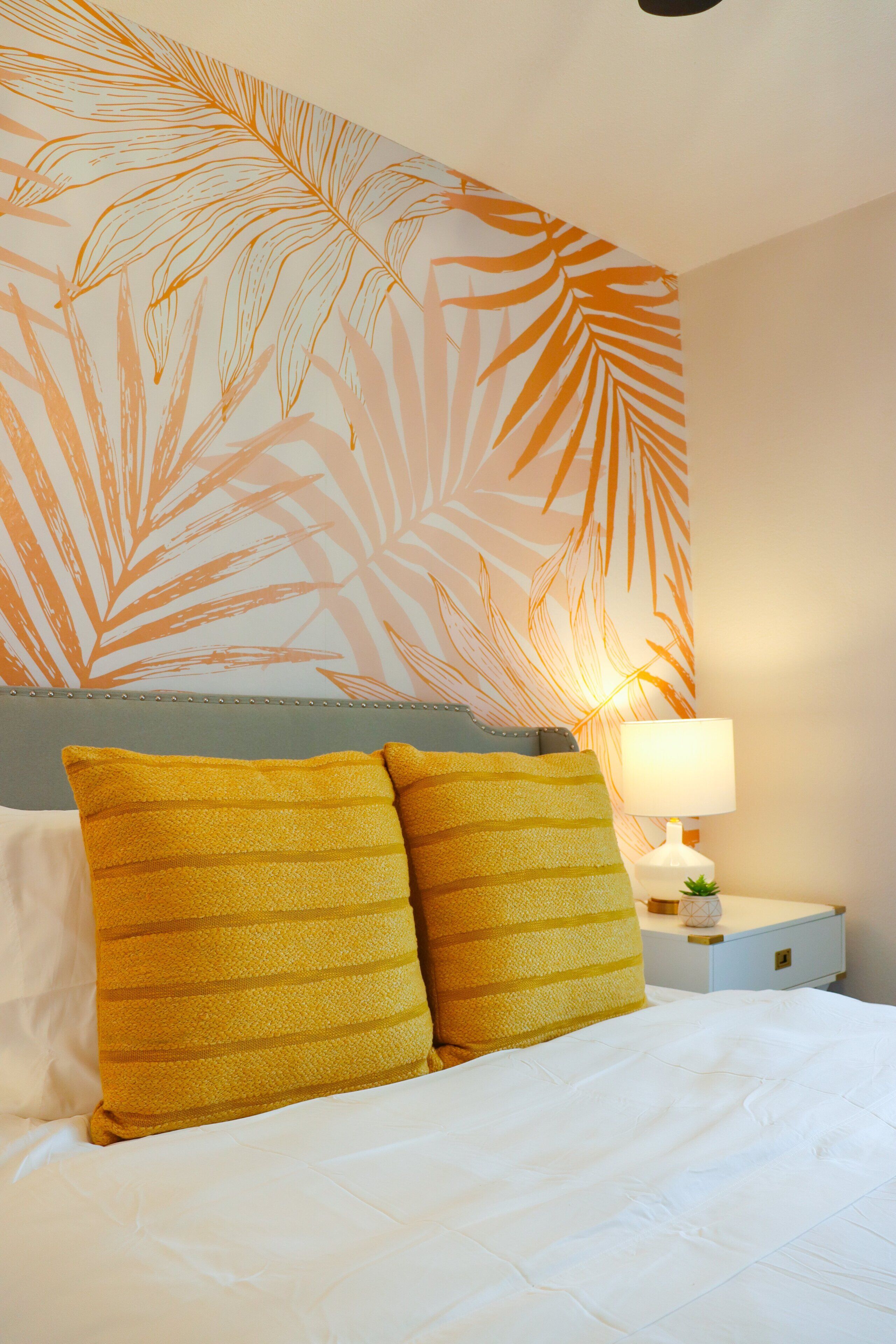 Une chambre d’hôtel avec un lit, deux oreillers, une table de chevet, une lampe et un mur orné d’un motif de feuilles.