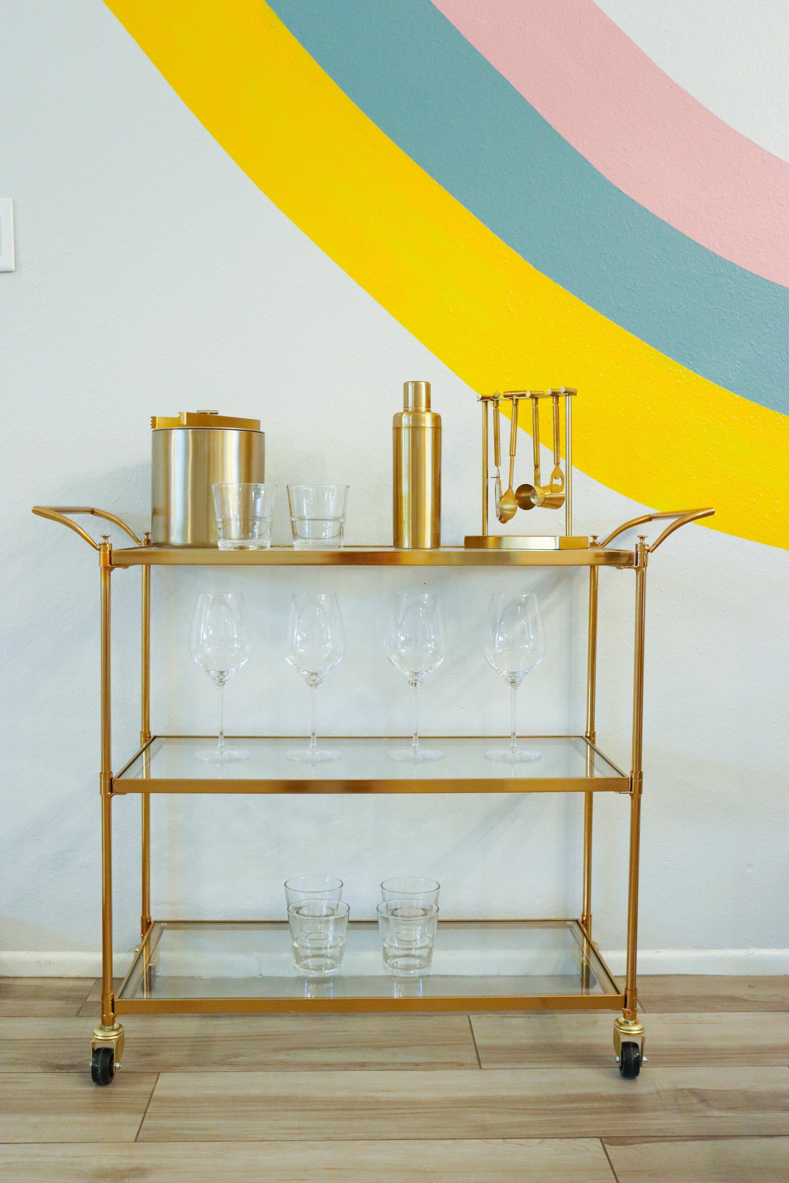 A gold-colored, wheeled cart with glass shelves holding wine glasses and a gold container.