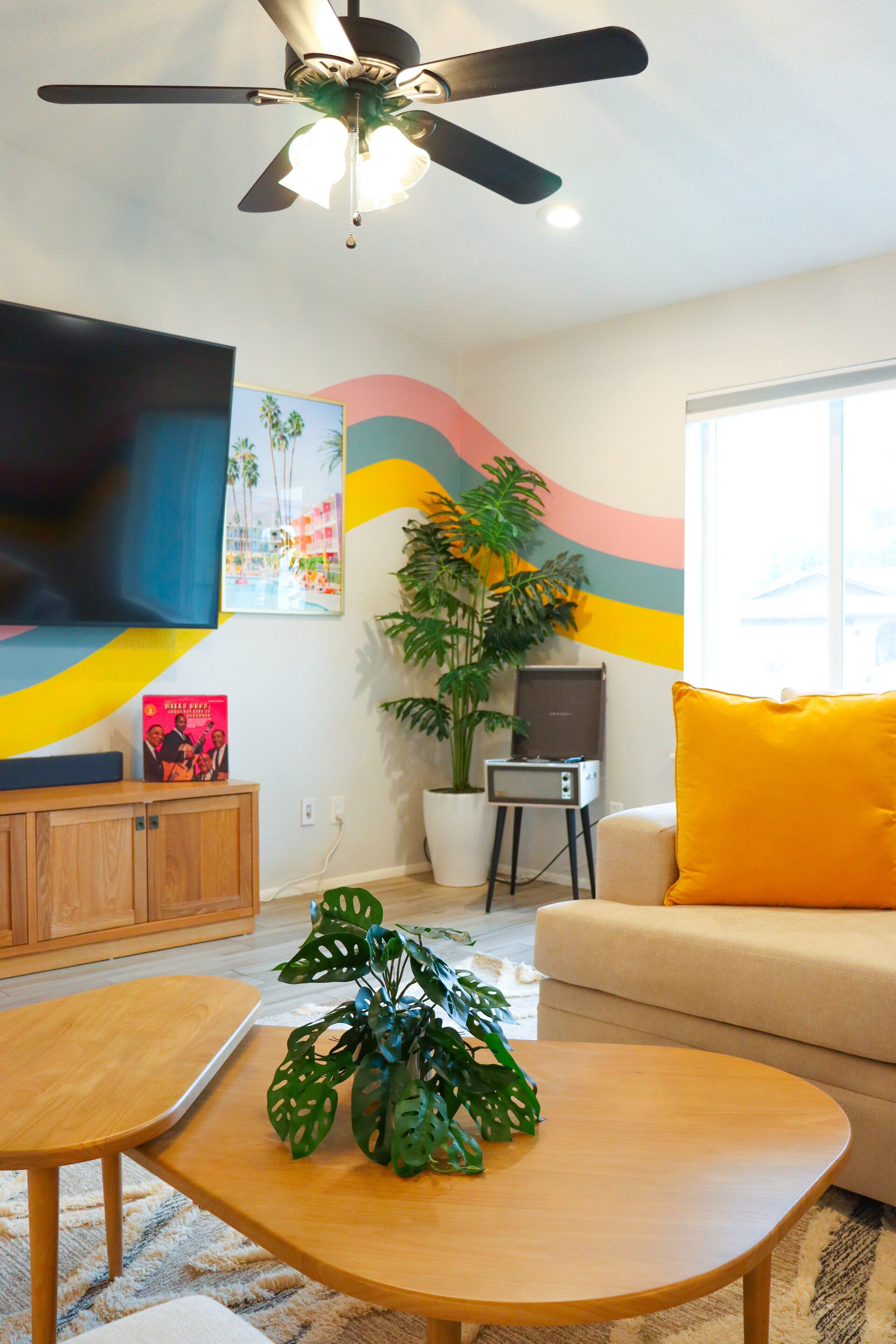 A living room with a ceiling fan, a flat-screen TV, a sofa with a yellow pillow, a wooden coffee table, and a potted plant.