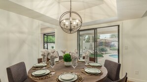 A round dining table with six chairs, set with plates, glasses, and napkins. A chandelier hangs above, and the room has large windows. - Beverly Scottsdale (Scottsdale)