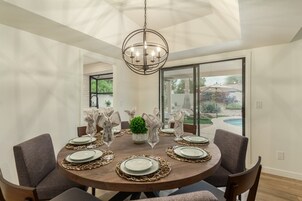 A round dining table with six chairs, set with plates, glasses, and napkins. A chandelier hangs above, and the room has large windows.