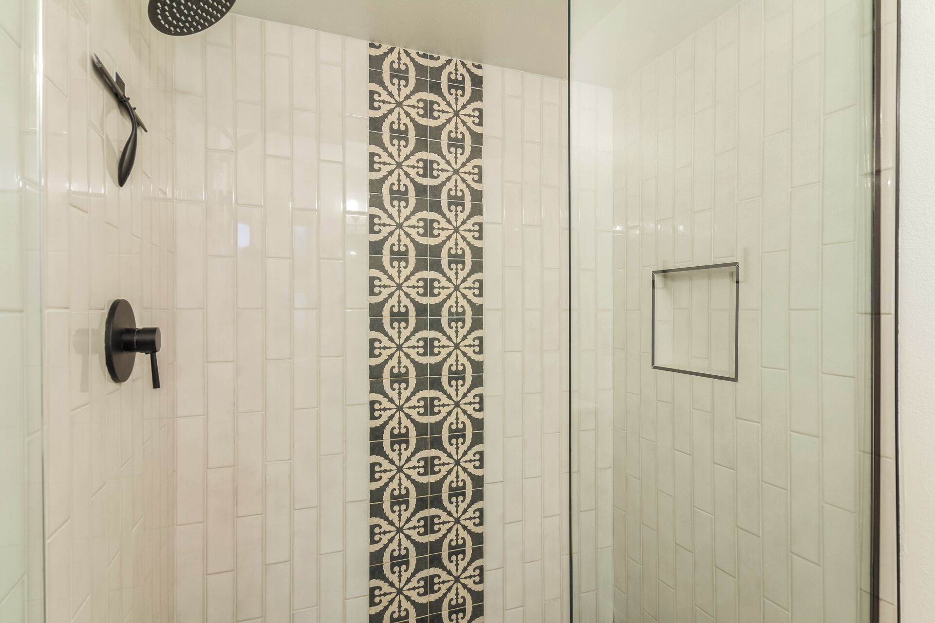 A shower with a patterned tile wall, a black showerhead, and a glass door.