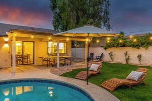 Un patio trasero con piscina, mobiliario exterior, un porche cubierto y luces colgantes.