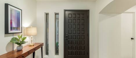 Un pasillo con una puerta de madera oscura, un cuadro enmarcado en la pared, una pequeña mesa con una lámpara y una planta, y suelo de madera.