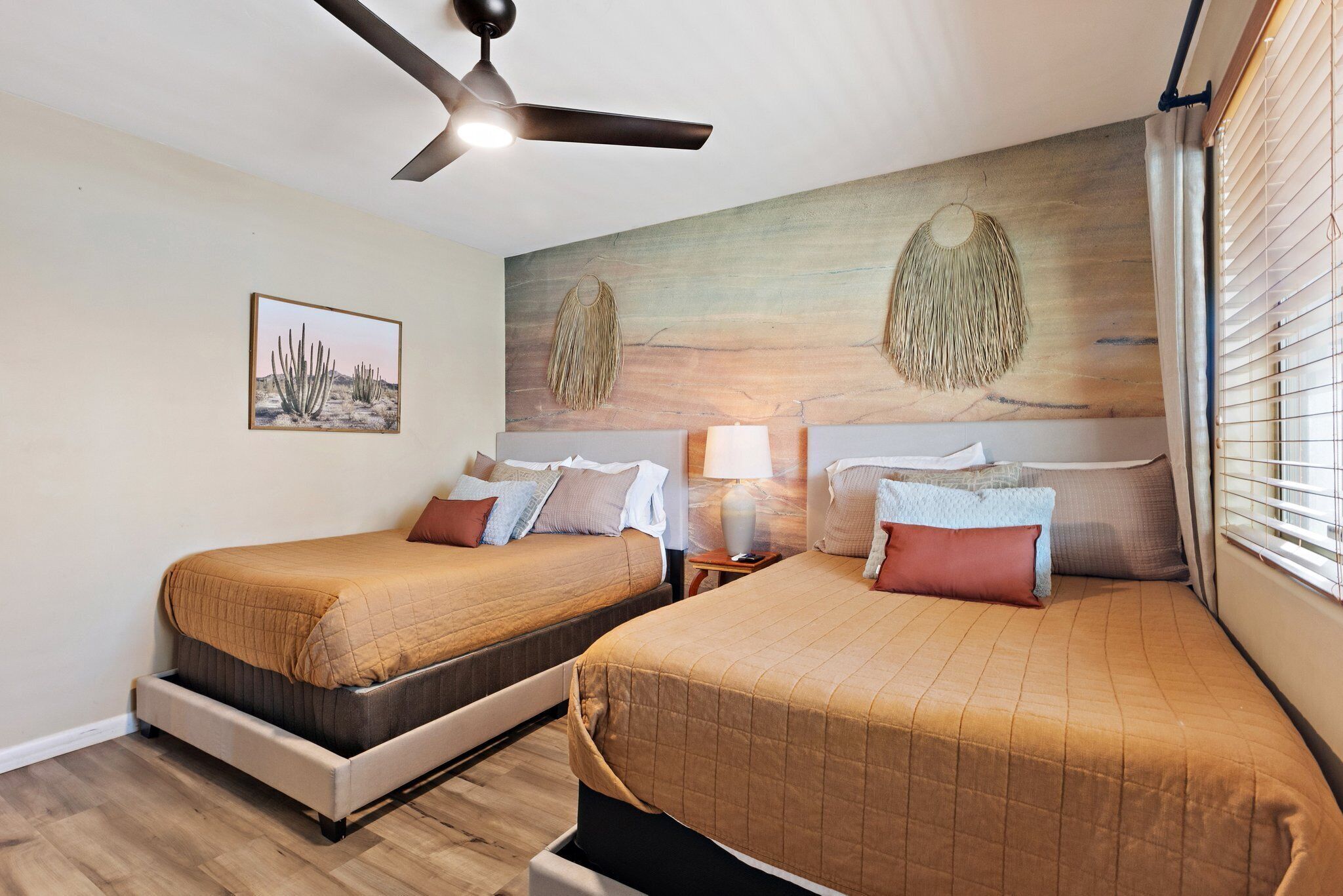 Una habitación de hotel con dos camas, un ventilador de techo, un cuadro con temática desértica y una lámpara de pared.
